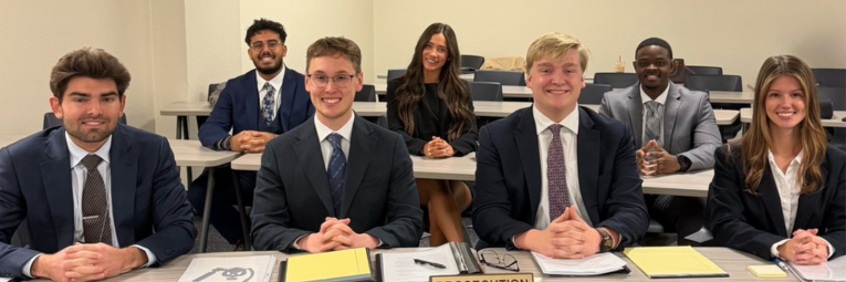 A group of students seated at a table in a classroom setting, dressed in formal attire.