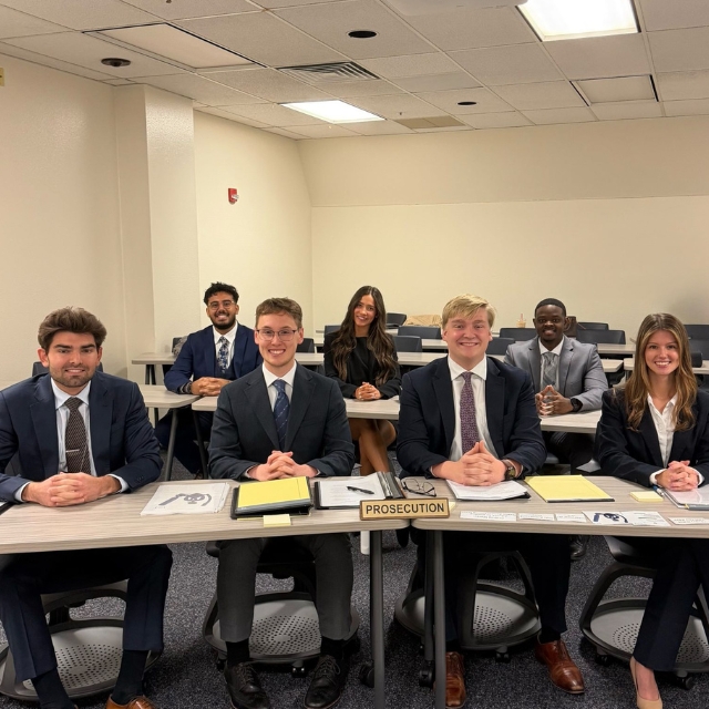 A grouStudents in formal attire sit at desks, with folders and documents labeled "Prosecution" in front of them. They are in a classroom setting, facing the camera, with a neutral wall in the background.