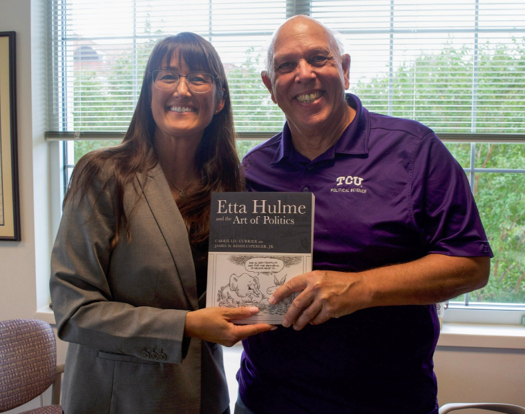 Carrie Currier and Jim Riddlesperger smile, standing indoors holding their book titled 