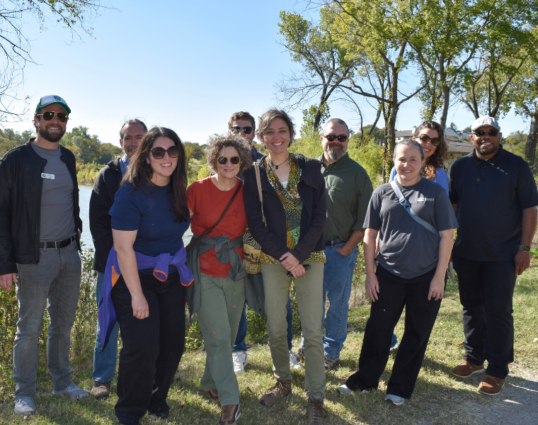 A group of TCU faculty smiling for a group picture at a site visit with a pond in the background.