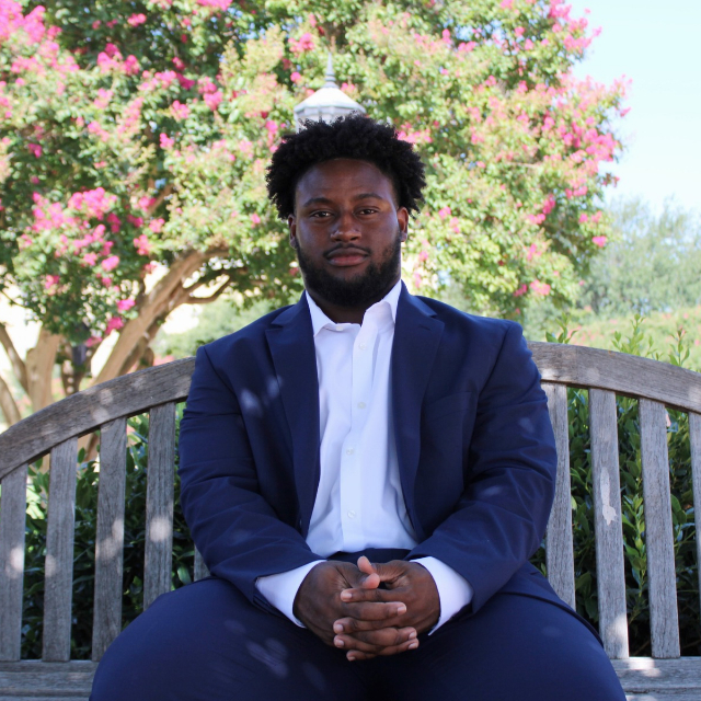 Isaac Shabay, in a blue suit, sits on a wooden bench in a garden. Pink flowering trees are in the background.