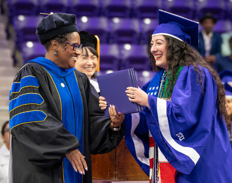 AddRan Dean Sonja Watson hands a diploma to a smiling Karina Torres wearing a purple cap and gown adorned with a Texas Christian University stole. They are in an indoor setting with purple seats in the background.